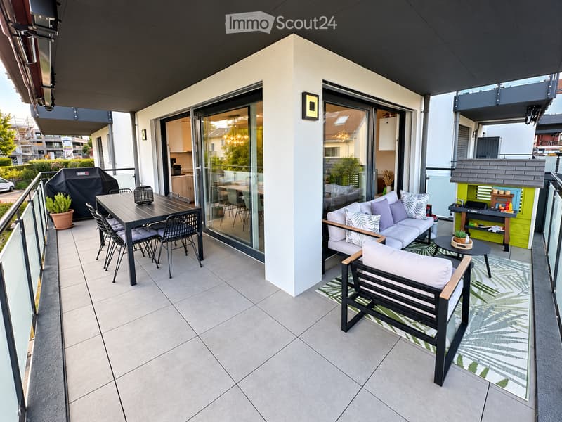 Modern balcony with grey sofa, white cushions, black bench, dining table, chairs, and a small grill. Features a green rug and potted plants.