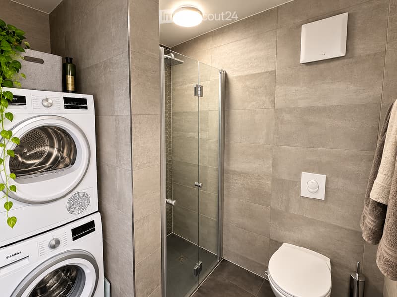 Modern bathroom with a glass-enclosed shower, a white toilet, a stacked washing machine, and a towel hanging on the wall.