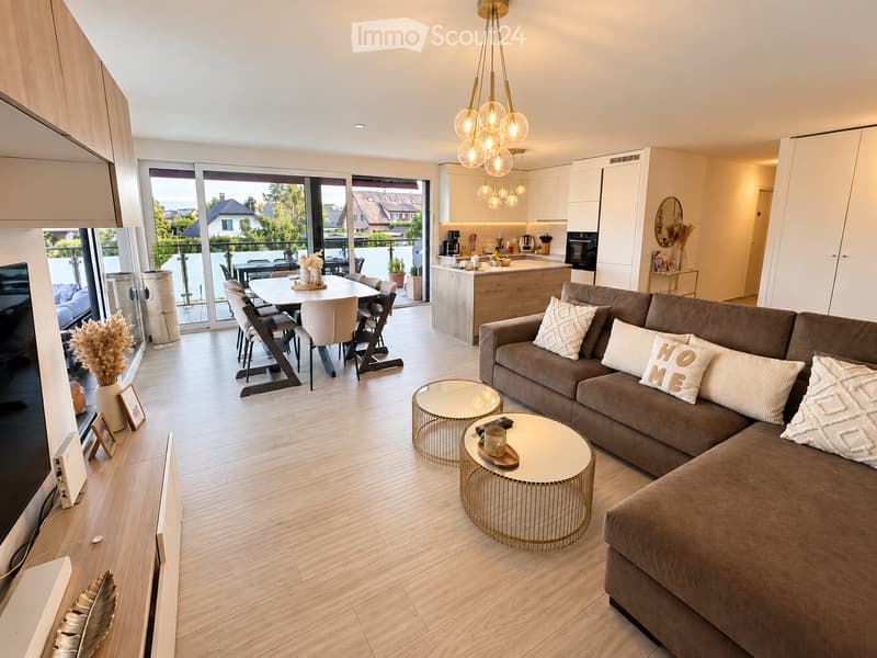 Modern living and dining area with a brown sectional sofa, coffee tables, dining table with chairs, and a kitchen area with cabinets and appliances. Large windows and sliding doors lead to a balcony.