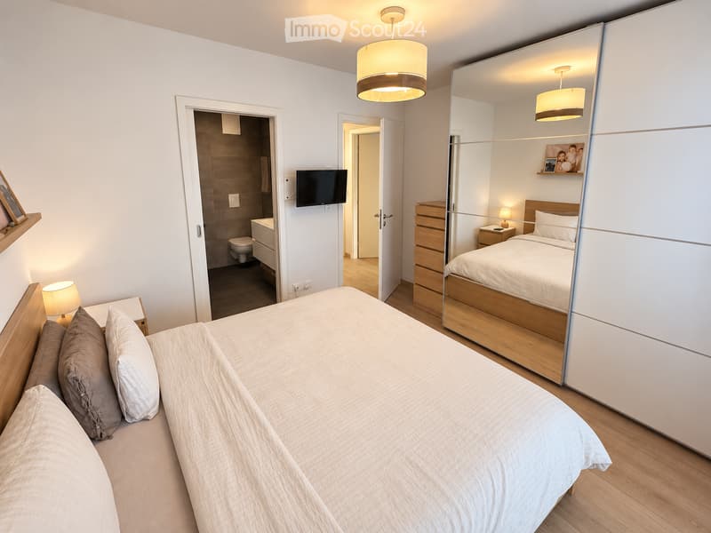 Modern bedroom with a large bed, nightstands, lamps, a large mirror, and a wardrobe. A television is mounted on the wall, and an adjacent bathroom is visible through an open doorway.