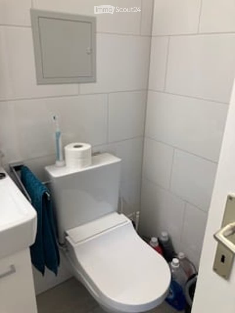 A compact bathroom with white tiled walls and floor, a toilet, a sink, a mirror, a towel, and cleaning supplies.