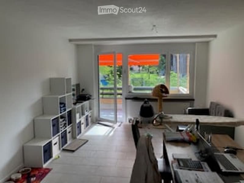 Bright, modern room with large window and sliding glass door leading to a balcony. Contains a desk with a laptop, chair, bookshelf, and a lamp.
