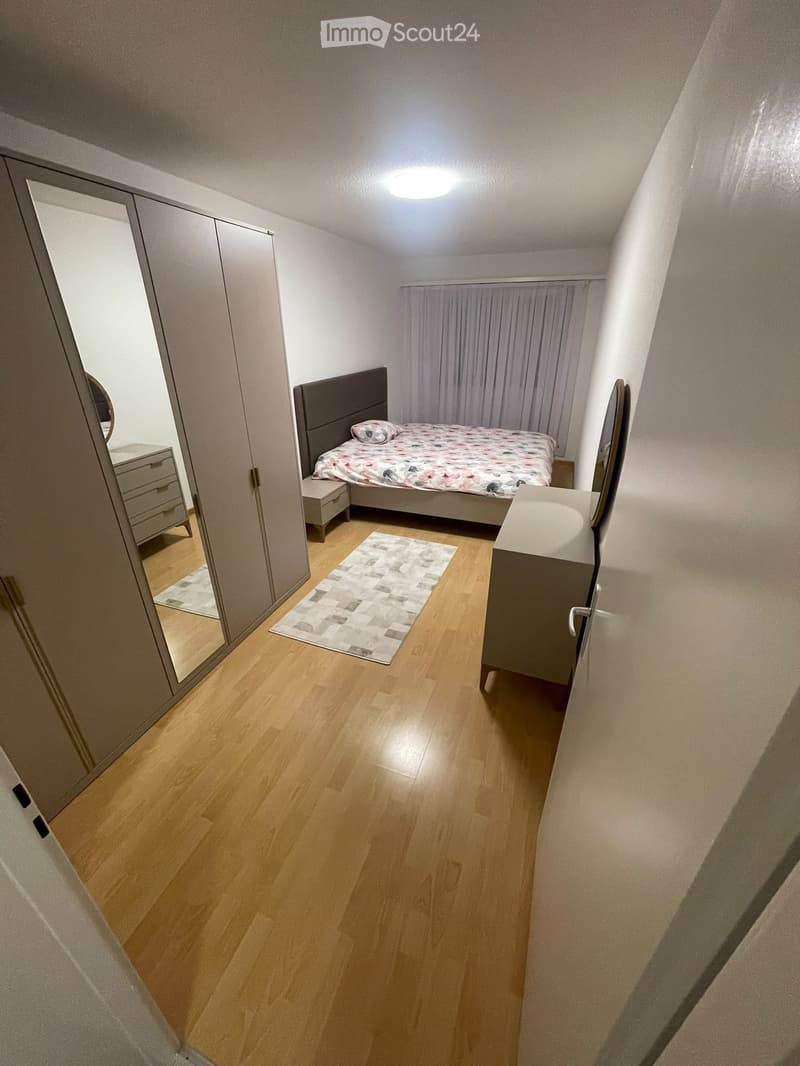 Modern bedroom with wooden flooring, a bed with a colorful bedspread, a nightstand, a wardrobe with mirrored doors, and a small rug in front of the bed.