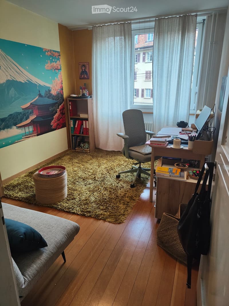 A well-furnished room with a sofa, desk, chair, bookshelf, laptop, and a large window with curtains. The room features a wooden floor, a shaggy rug, and a decorative painting on the wall.