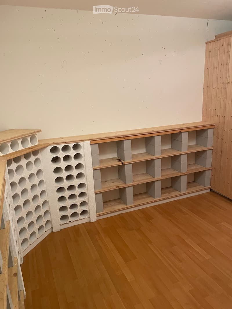 Empty room with wooden flooring and white walls, featuring a built-in wooden shelf unit with multiple levels and a wine rack section.