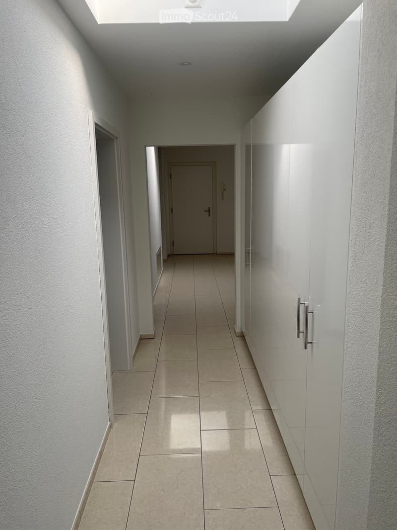 Clean, modern hallway with tiled floor, white walls, and recessed lighting. Features a closed door at the end and built-in cabinetry on one side.