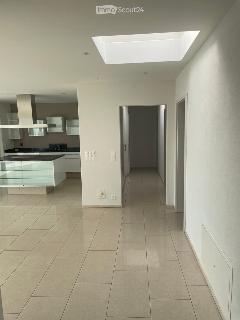 Empty hallway with tiled floor, white walls, and a skylight at the top. The hallway leads to a kitchen area with white cabinets and a black countertop.