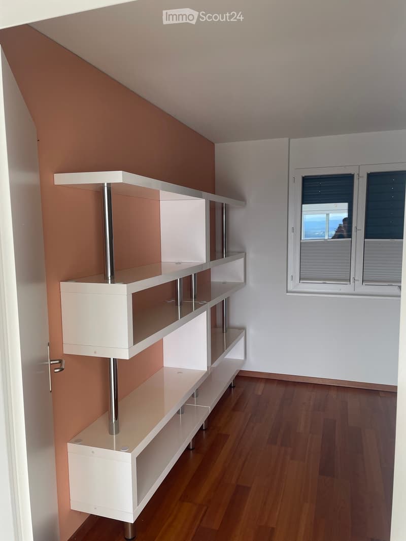 Empty room with wooden flooring, white walls, and a modern white shelving unit with glass shelves and metal supports. A window with blinds offers a view outside.