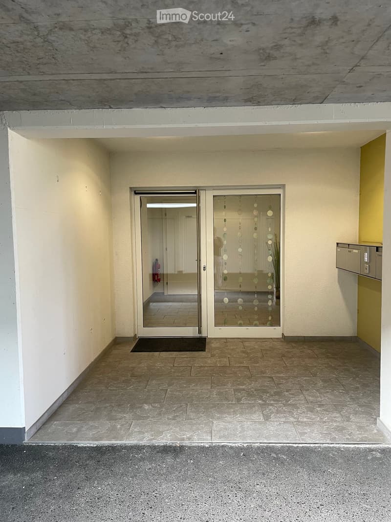 Empty hallway with tiled floor, glass double doors, and a small mat. The walls are painted in neutral tones, and there is a small utility area visible on the right side.
