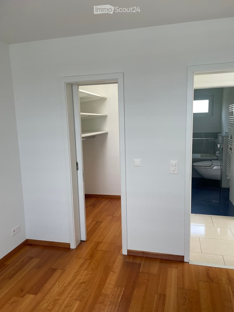 Empty hallway with light wooden flooring, white walls, and two open doors leading to a closet and a bathroom.