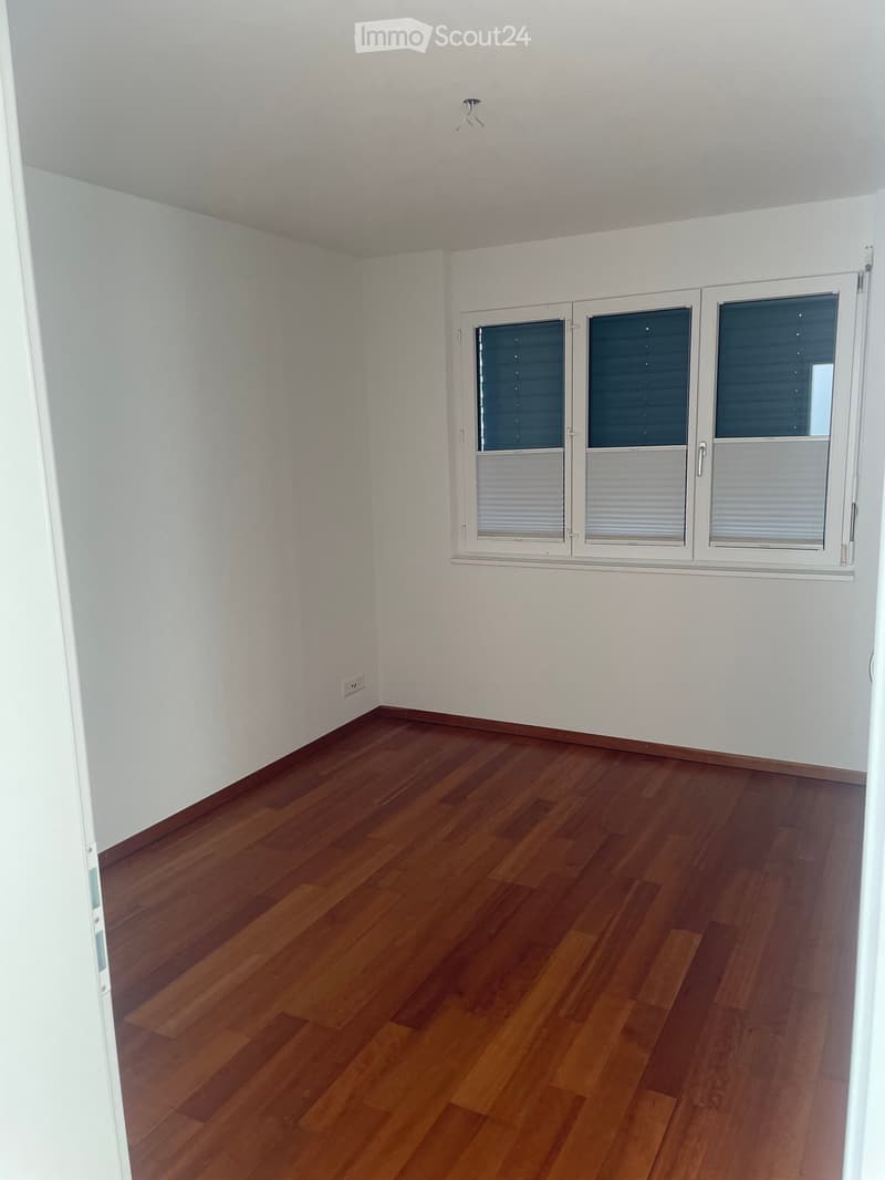 Empty room with wooden flooring and white walls, featuring a large window with blinds and a visible electrical outlet on the wall.