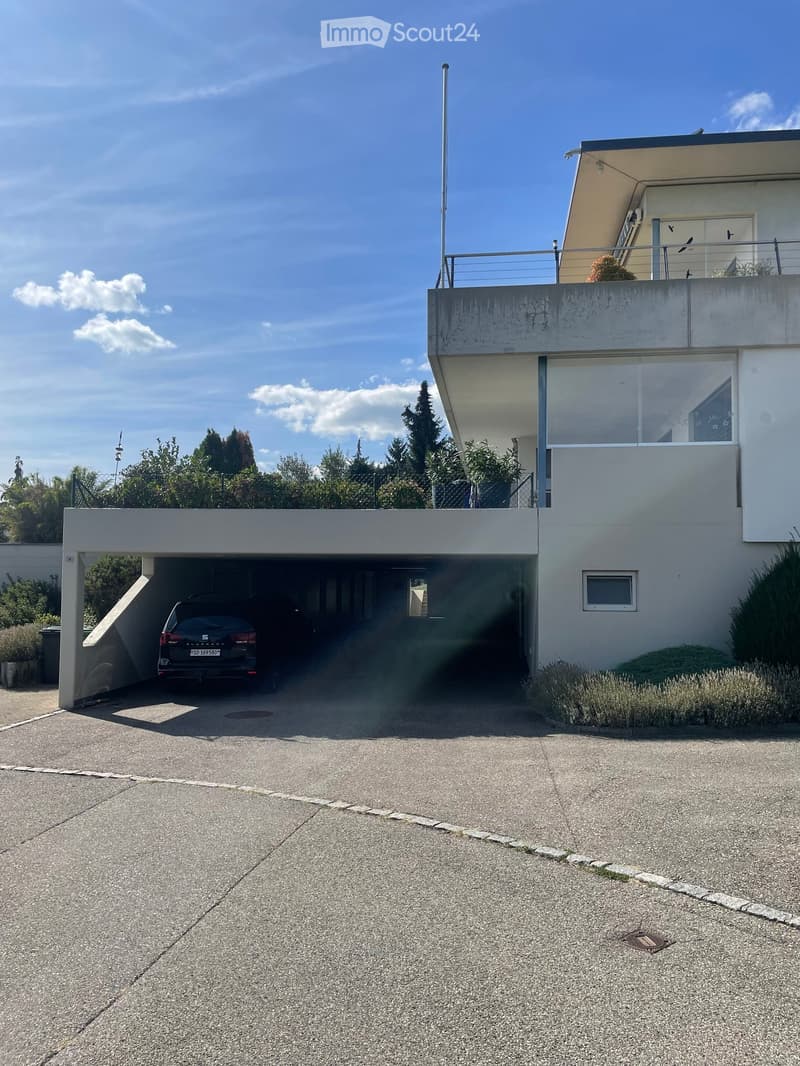 Modern two-story house with a raised ground floor, garage, and balcony. The garage has a car parked inside, and the house features a sleek, contemporary design with large windows and a flat roof.
