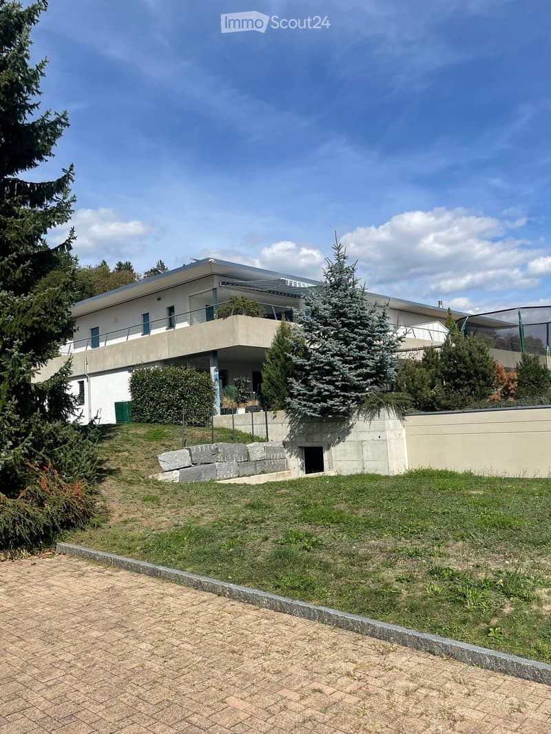 Modern two-story house with white walls, large windows, and a flat roof, surrounded by greenery and a raised ground floor.