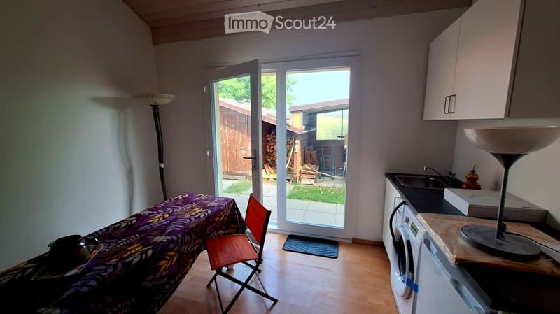 A small, furnished room with a wooden floor, a covered table with a chair, a washing machine, a sink, and a floor lamp. A sliding glass door leads to a backyard.