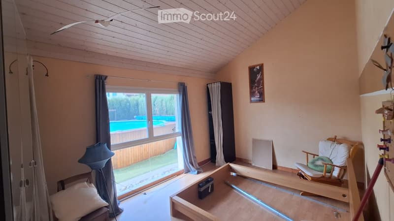 Bright room with wooden ceiling, large windows, and a view of a swimming pool. Contains a wooden chair, a lamp, and a picture frame on the wall.