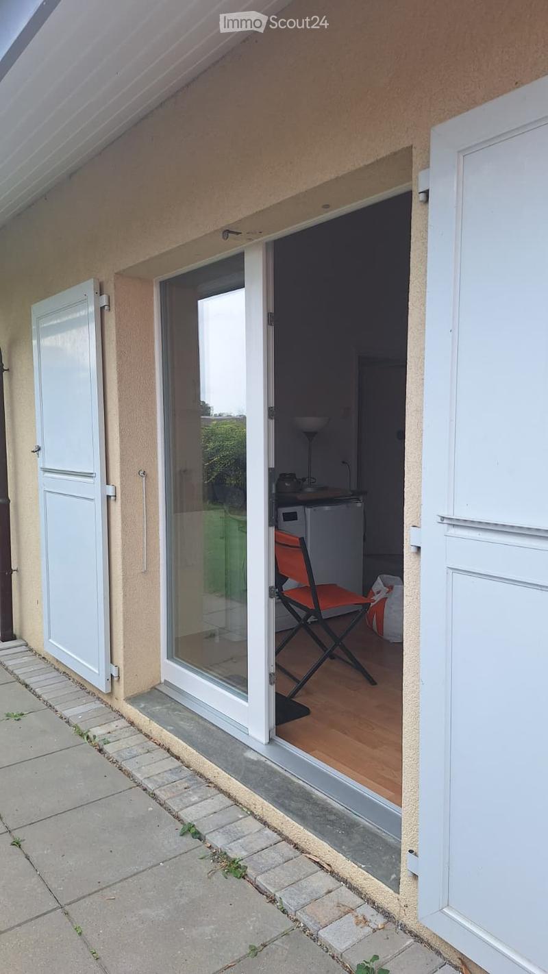 Balcony with glass sliding doors leading to a small indoor space, featuring a wooden floor, a red folding chair, and a small table with a lamp.