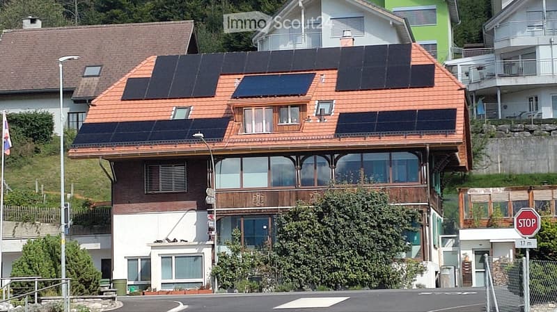 Two-story house with solar panels on the roof, large windows, and a balcony. The house is surrounded by greenery and other residential buildings.