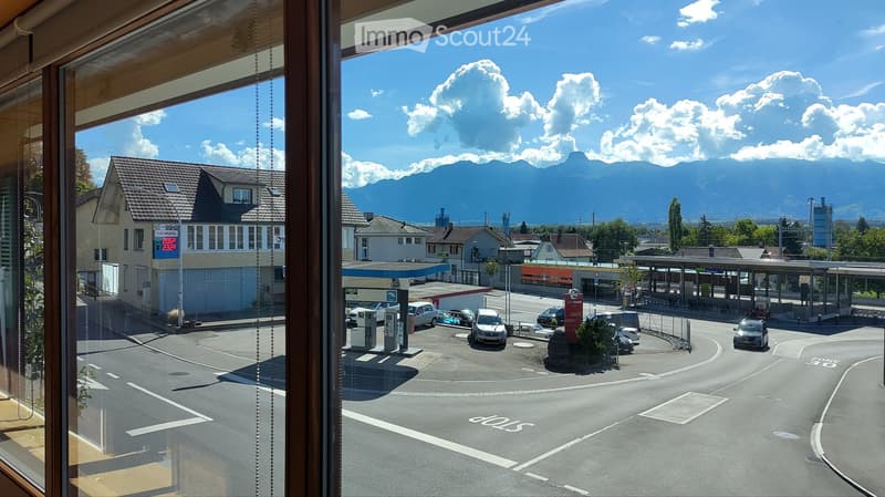 View from a window showing a town square with a gas station, parked cars, and distant mountains under a clear blue sky.
