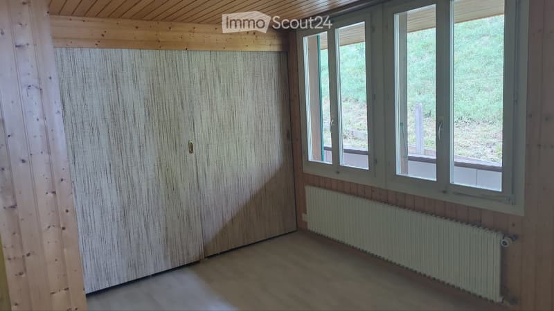 Empty room with wooden walls, ceiling, and floor, large windows, radiator, and a door.