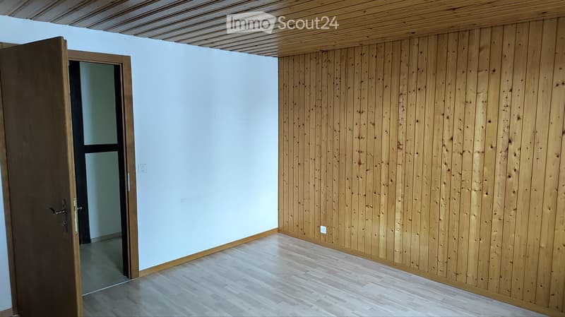 Empty room with light wooden walls and floor, white wall on one side, and a partially open door leading to another space.