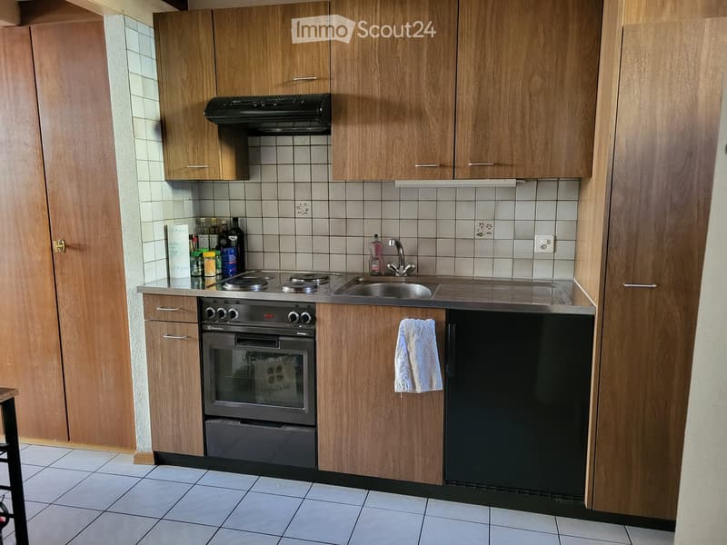 Compact kitchen with wooden cabinets, stainless steel sink, oven, and refrigerator. Countertop holds various items including bottles and a towel.