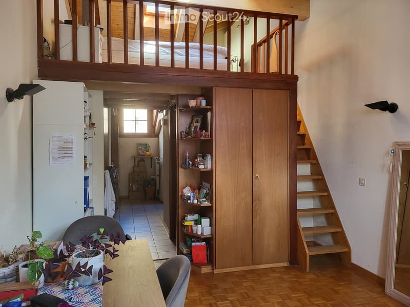 The image shows a cozy, multi-level living space with a dining area on the ground floor and a lofted bedroom area above. The ground floor features a wooden dining table with chairs, potted plants, and a cabinet with shelves. A staircase leads up to the lo