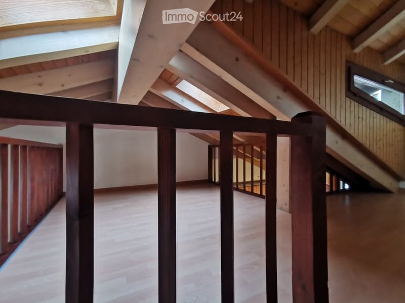 Empty room with wooden flooring and exposed wooden beams on the ceiling. The room has large windows allowing natural light to enter, and a staircase leading to another level.