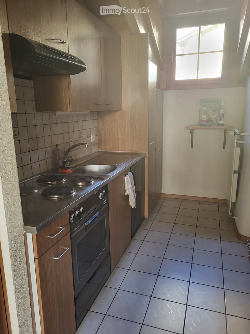 Modern kitchen with wooden cabinets, tiled floor, and stainless steel appliances including an oven, sink, and faucet. A small shelf holds a few items, and a towel hangs from a rack.