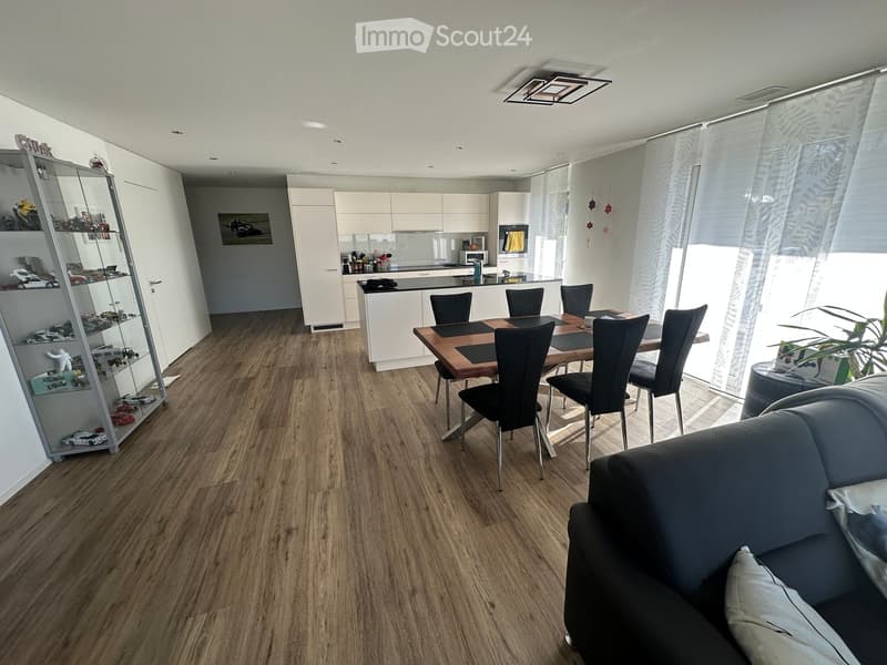 Open-plan living and dining area with wooden flooring, a grey sofa, a dining table with chairs, a kitchen area with cabinets and appliances, and a display shelf with model cars.