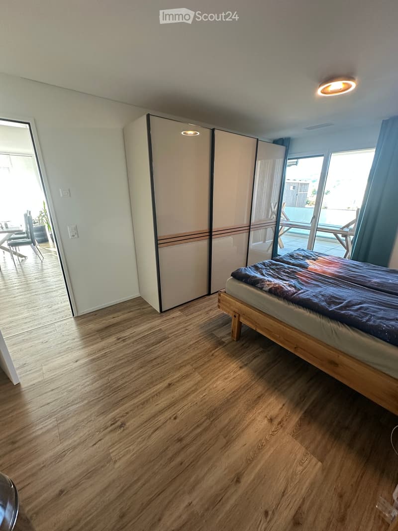 Bright bedroom with wooden flooring, a bed with a colorful duvet, a large wardrobe, and a view of a patio area with outdoor furniture.
