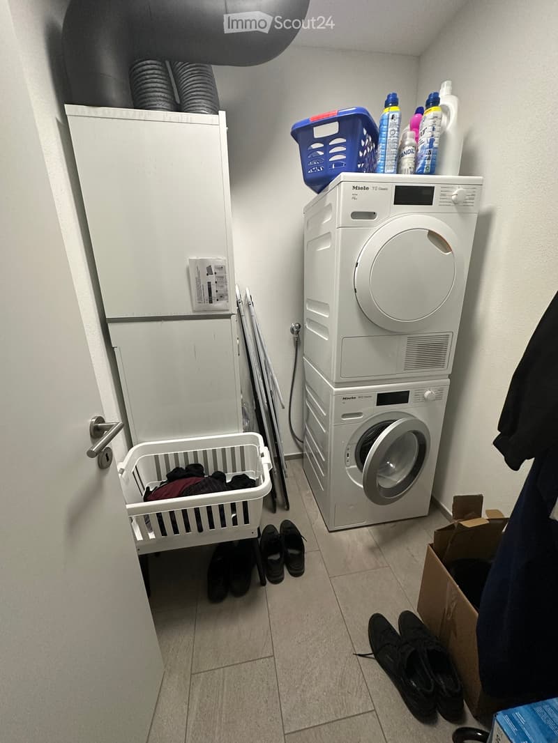 Laundry area with a white washing machine, a laundry basket, cleaning supplies, and shoes on the floor.
