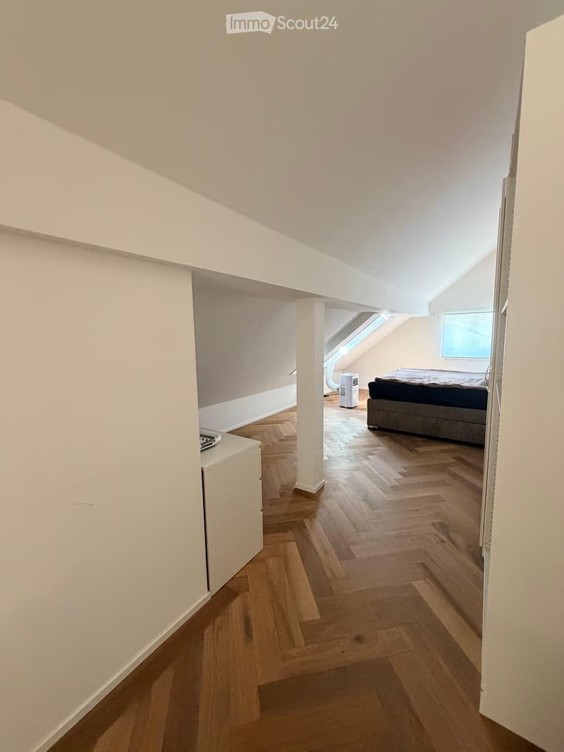 A modern, well-lit bedroom with wooden herringbone flooring, white walls, and a large window. Features a bed, a small cabinet, and an air conditioner unit.