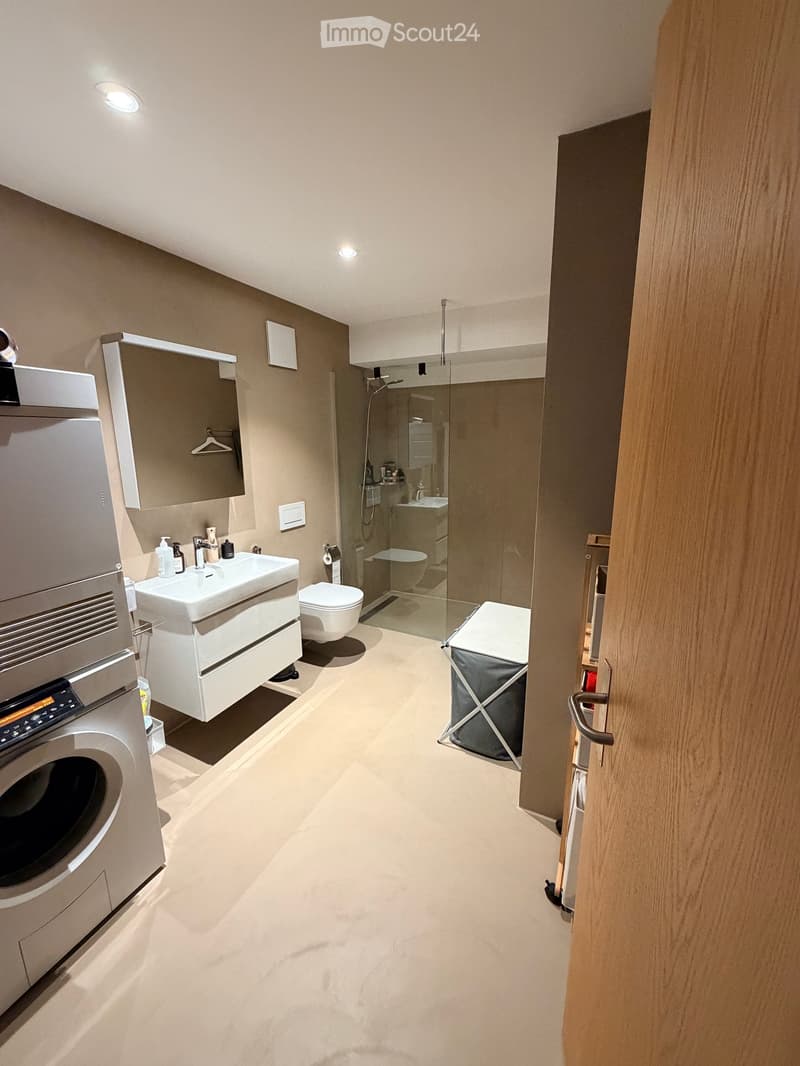 Modern bathroom with a white sink, toilet, glass-enclosed shower, and a washing machine. Features a large mirror, built-in cabinet, and a laundry basket.