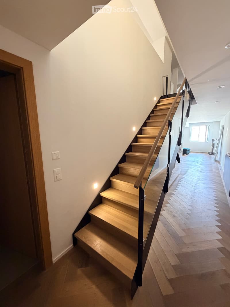 Modern staircase with wooden steps and a glass railing, leading to an open area with wooden flooring and a visible room in the background.
