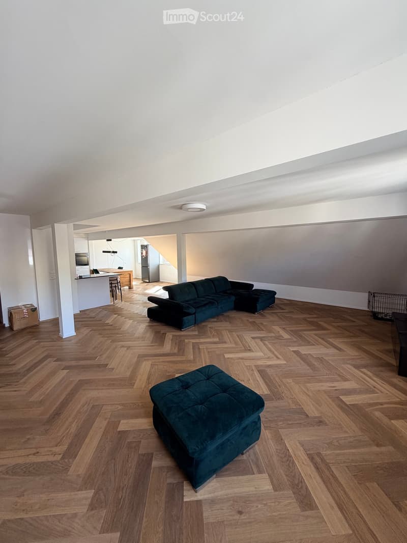 Open-plan living and dining area with wooden herringbone flooring, a dark green sectional sofa, ottoman, and a small dining area with a table and chairs. A modern kitchen is visible in the background with white cabinets and stainless steel appliances.