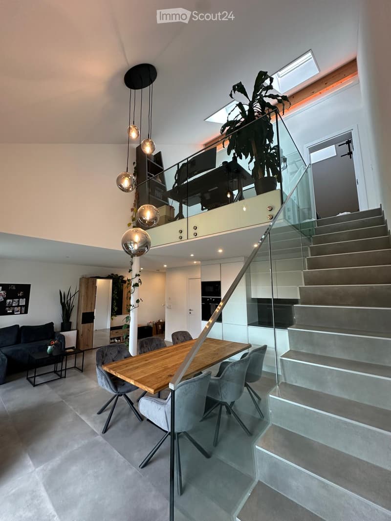 Modern open-plan living and dining area with grey sofa, wooden dining table, grey chairs, pendant lights, and a staircase leading to an upper level.