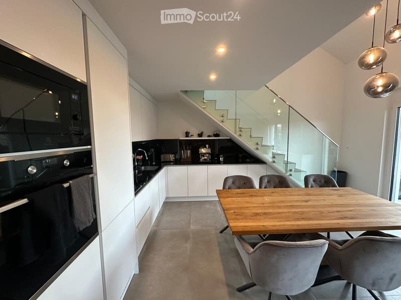 Modern kitchen with white cabinets, a wooden dining table, four chairs, a built-in oven, and a staircase leading to another level.