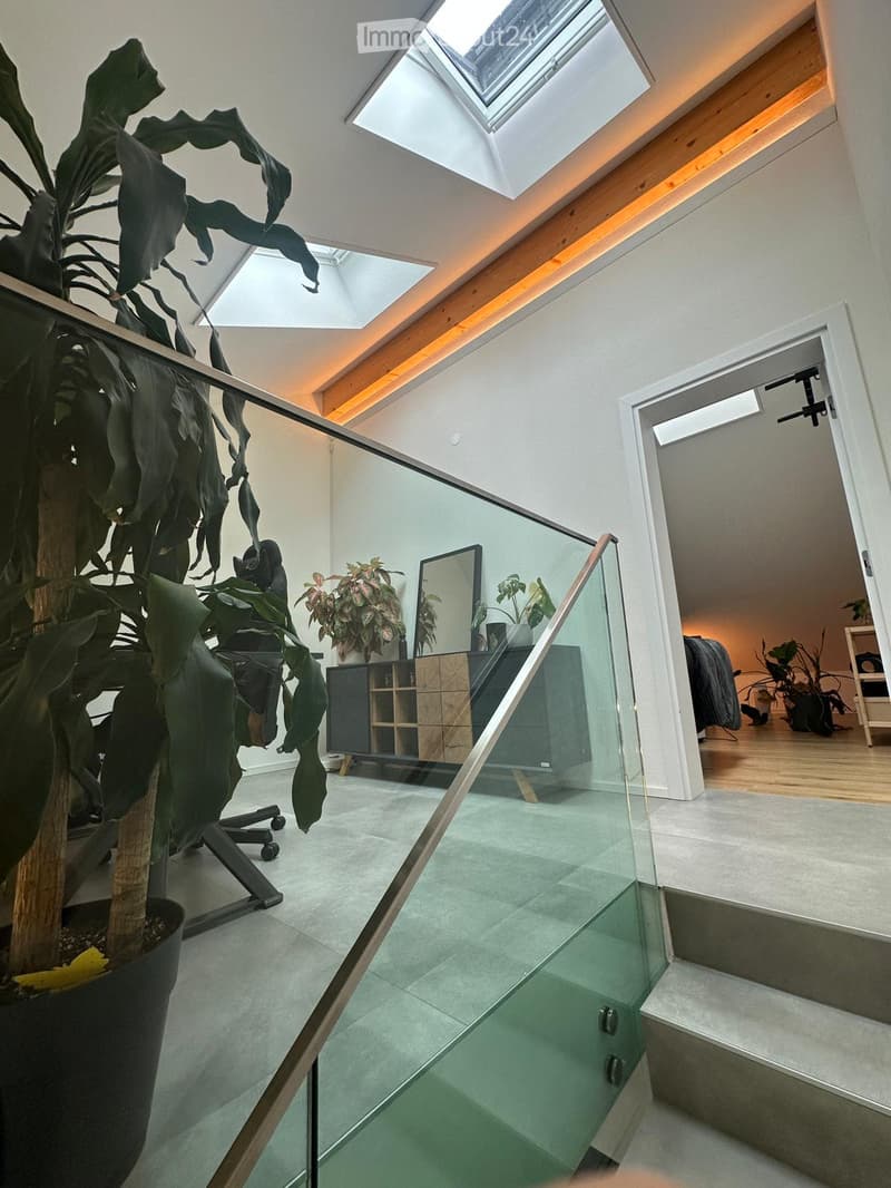 Modern, well-lit room with a glass staircase leading to another level, featuring a large potted plant, a chair, a cabinet, and a mirror.