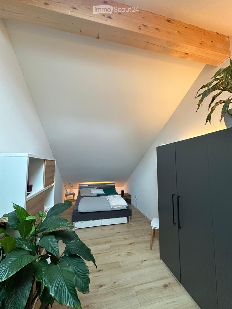 Modern bedroom with wooden floor, slanted ceiling, bed with green bedding, nightstand, wardrobe, and plants.