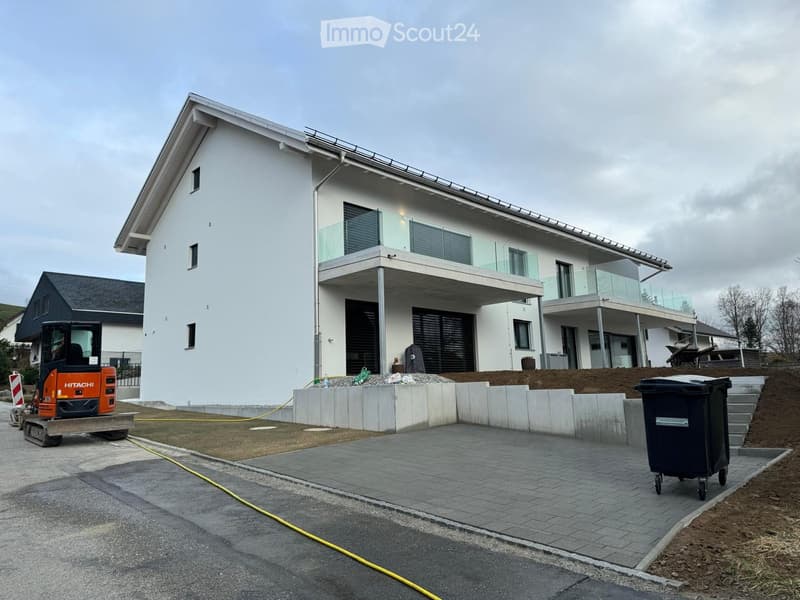Modern two-story house with white exterior, large windows, and a raised ground floor. Features include a balcony, garage, and construction equipment nearby.