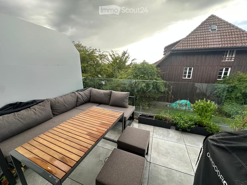 Modern balcony with grey sectional sofa, wooden coffee table, and ottomans, surrounded by glass railing and greenery.