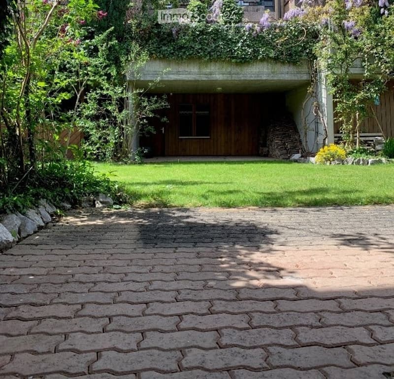 Garden area with a paved brick pathway leading to a house entrance, surrounded by greenery and flowers.