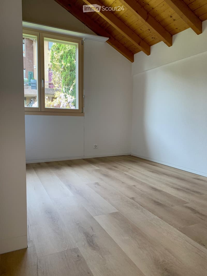 Empty room with light wooden flooring, white walls, and a large window with a view of greenery outside.