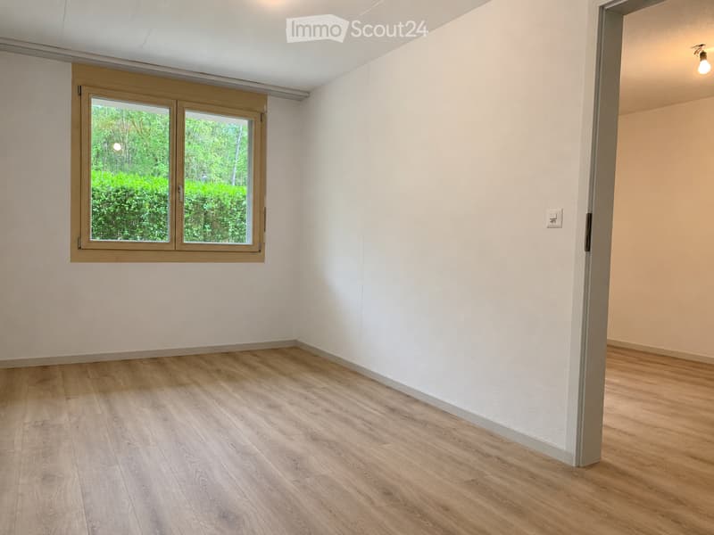 Empty room with light wooden flooring, white walls, and a large window with a view of greenery.