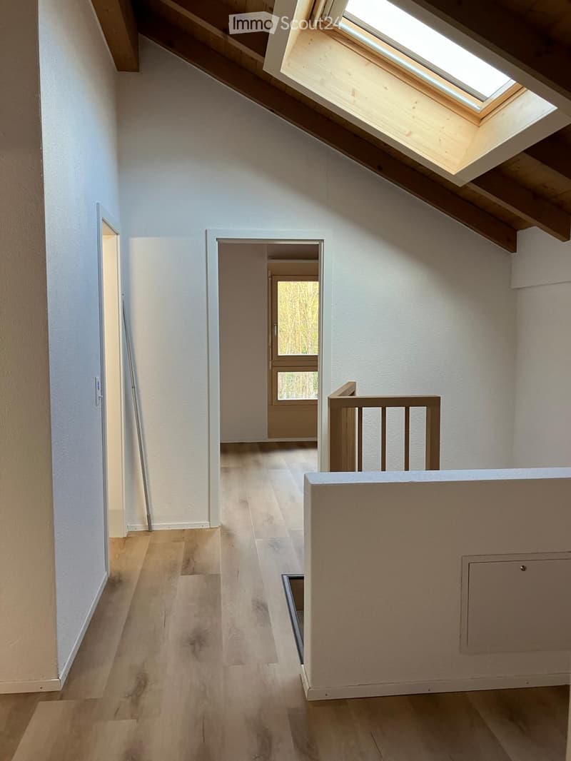 Bright hallway with wooden flooring, white walls, and a skylight at the top. Features a staircase railing and an open doorway leading to another room.
