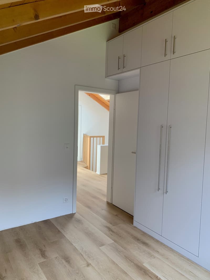 Empty room with light-colored wooden flooring and white built-in cabinets with silver handles. An open doorway leads to another room with wooden flooring and a visible staircase.