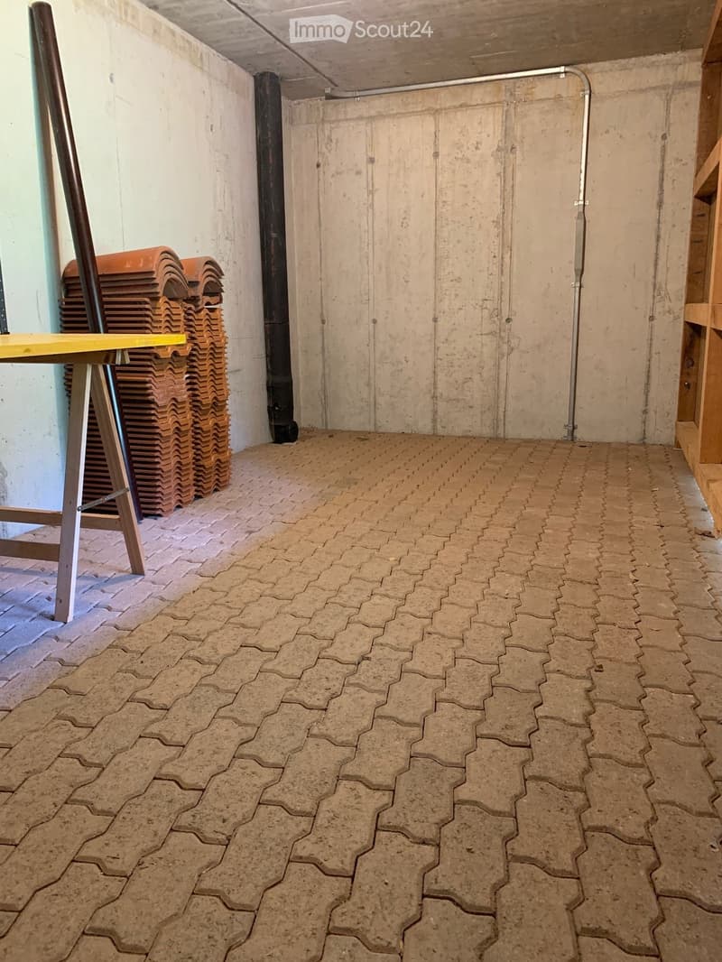 Empty room with concrete walls and a patterned tile floor. A stack of orange plastic chairs and a wooden table are visible on the left side.