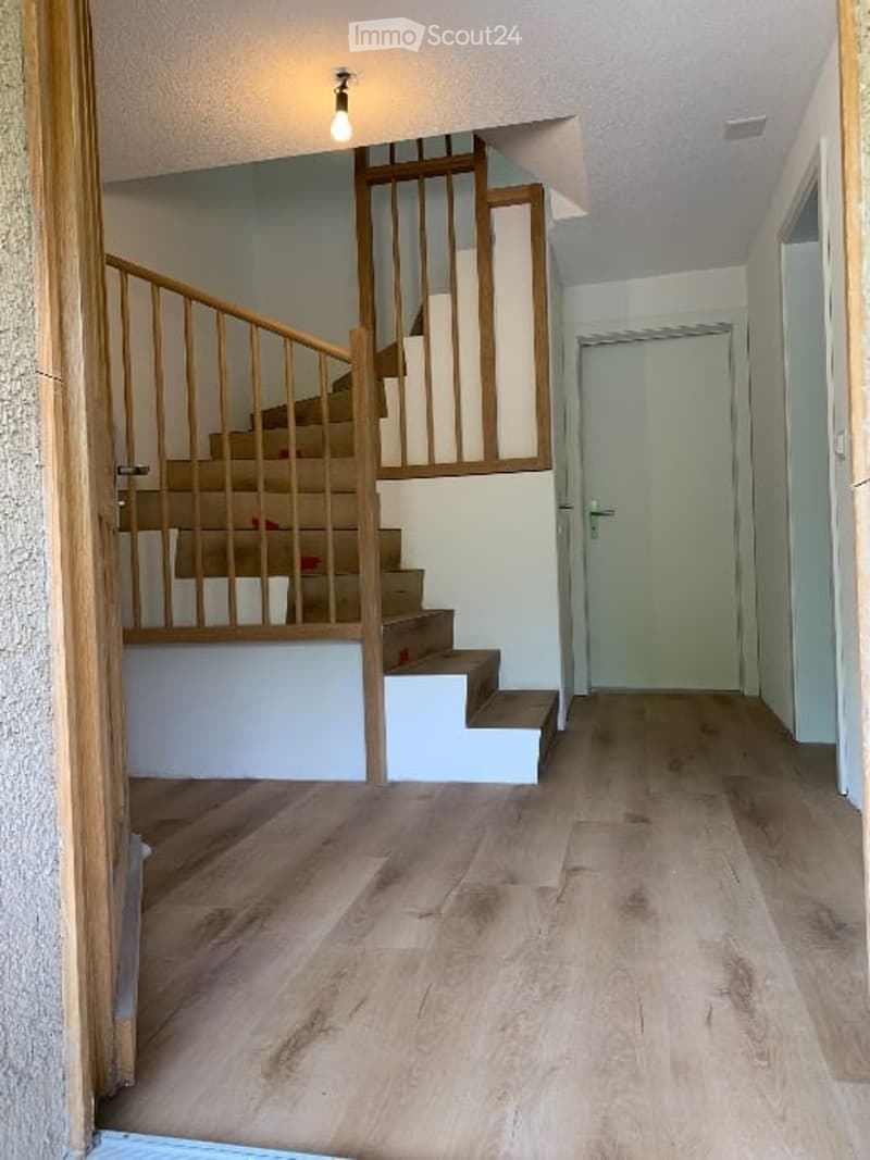 Spacious hallway with wooden flooring, a staircase with wooden railings, and a closed door leading to another room.