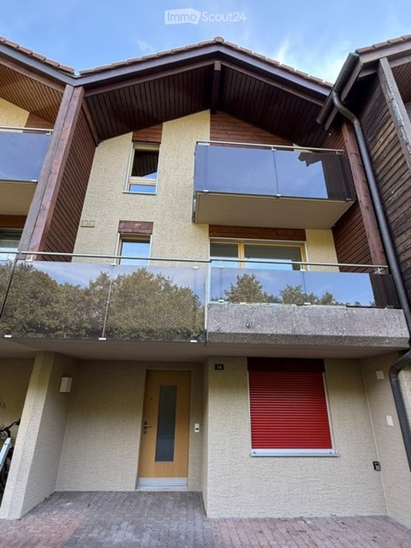 Modern two-story building with a sloped wooden roof, large glass balconies, and a red shuttered window on the ground floor.