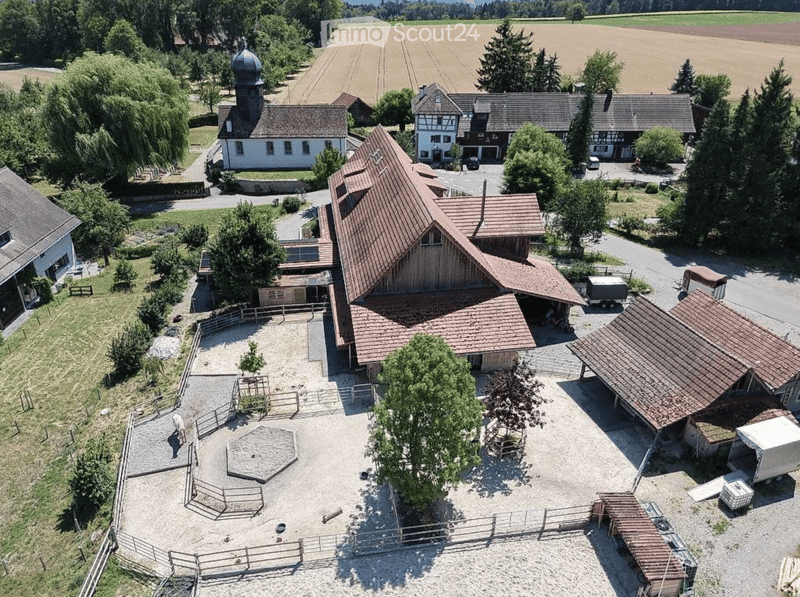 Aerial view of a rural estate with multiple buildings, including a large barn, residential houses, and a fenced area with a circular sandy arena.